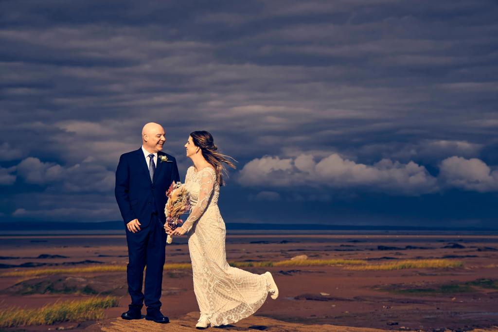 Bride and groom embracing on a coastal landscape beneath dramatic evening clouds