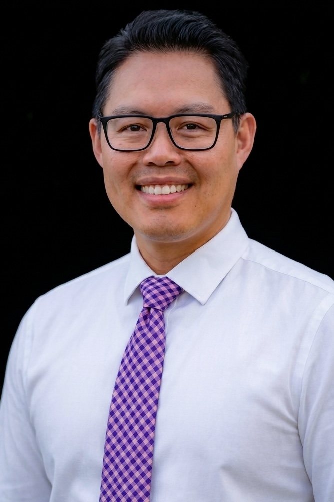 Professional studio headshot of a man wearing a white shirt and patterned purple tie.